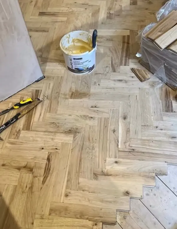 Herringbone wood floor being laid, with a tub of adhesive and offcuts on the boards